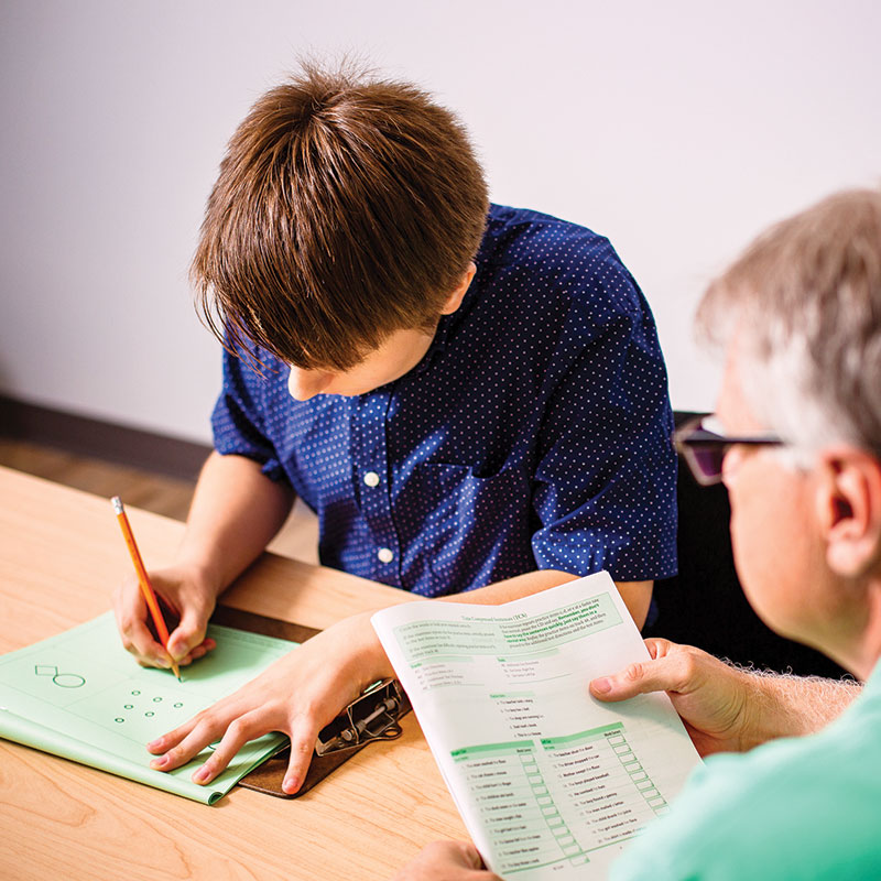 A client sits at a table filling taking a writing test with a clinician on looking during a neuropsychological test.