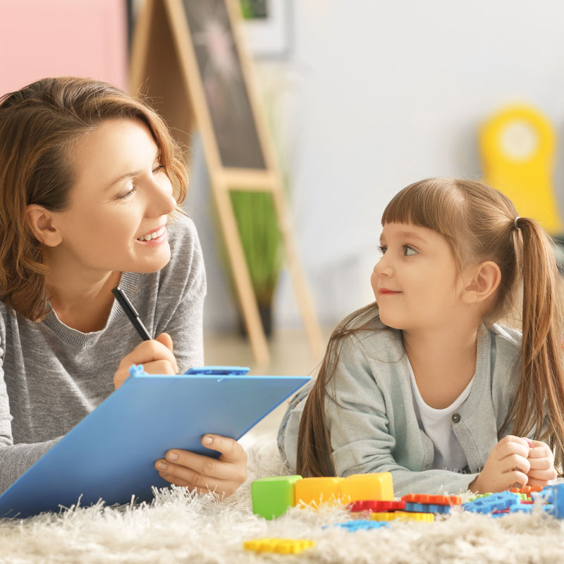 A therapist with a clipboard talks with a child on the floor demonstrating psychological testing. 